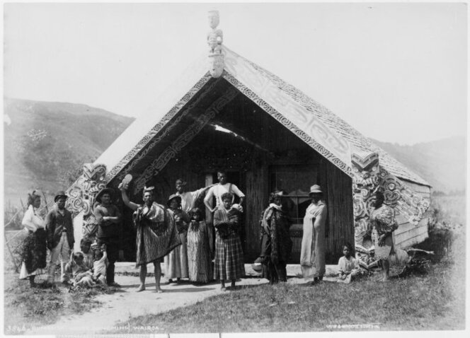 Photograph of an unidentified Māori group in front of the Hinemihi meeting house at Te Wairoa. Taken by Burton Bros in 1880s and before 10 Jun 1886 when it was buried in the Mount Tarawera eruption.