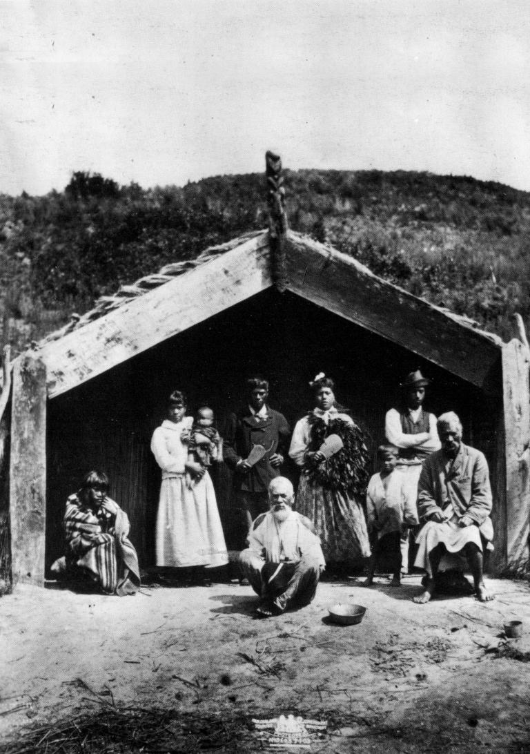 Photograph showing a Māori group of men, women, and children, outside a whare in the Rotorua district. The two seated men were guides on the pink and white terraces. Photograph taken between 1871 and 1886 by Elizabeth Pulman. Note on back of print reads: "The two old Maoris sitting down guided on the pink and white terraces. The whole party and place were destroyed."