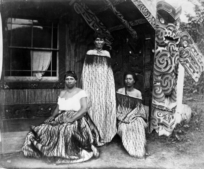 Tourist guides Sophia Hinerangi (standing), Kate Middlemass (Kati) (on left) and another guide, outside Hinemihi meeting house at Te Wairoa. Photograph by Elizabeth Pulman around 1881.